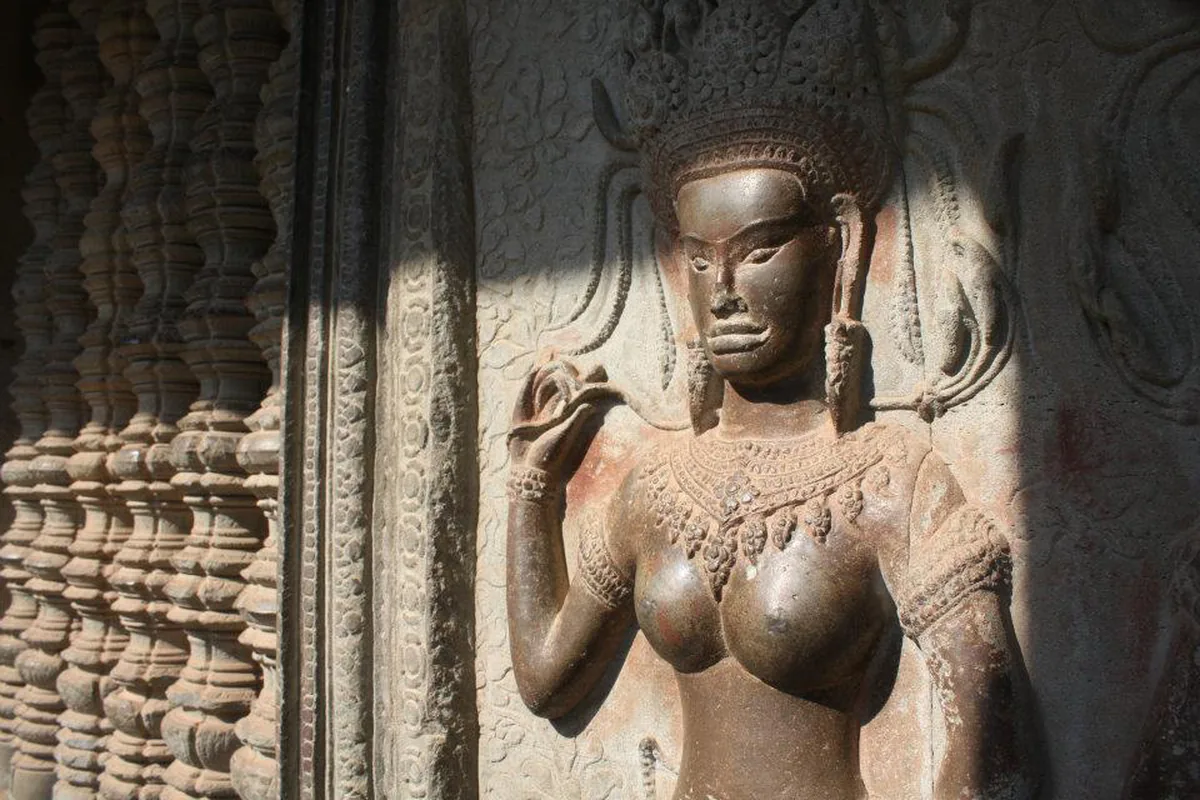 Apsara vytesaná v kameni na reliéfu v Angkor Wat, ve vysoké pokrývce hlavy se zdviženou rukou, vedle sloupu ze soustruženého kamene.