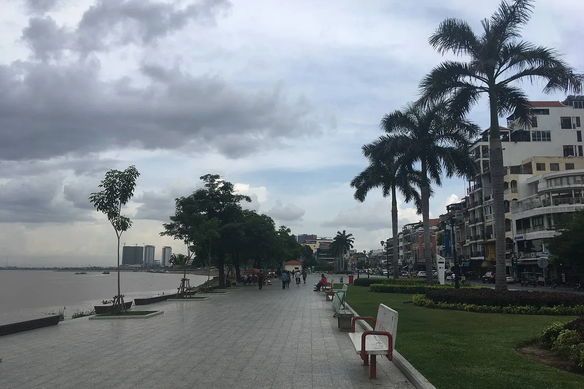 Dlážděná promenáda vedle Tonlé Sap v Phnom Penhu pod zataženou oblohou, s královskými palmami a budovami vpravo a řekou vlevo.
