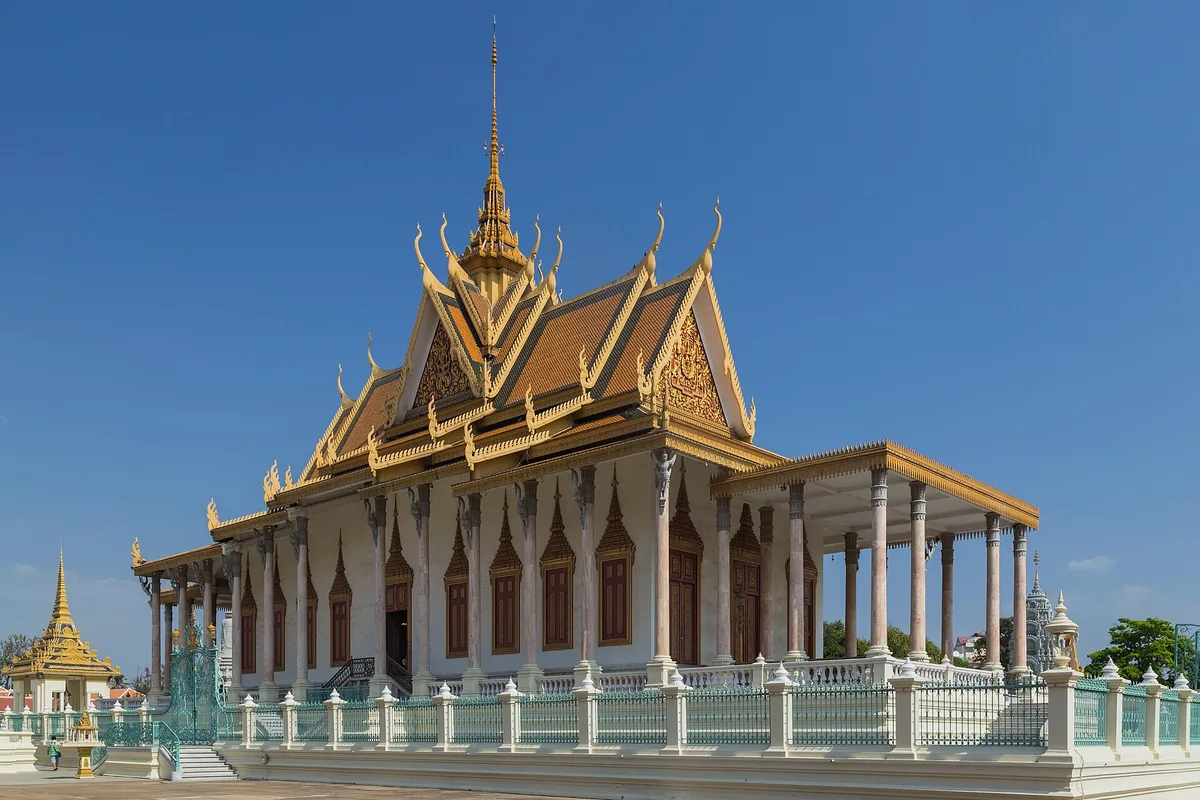 Stříbrná pagoda v Phnom Penhu, bílý sál se zlatými stupňovitými střechami a vysokou věží, za bleděmodrým zábradlím.