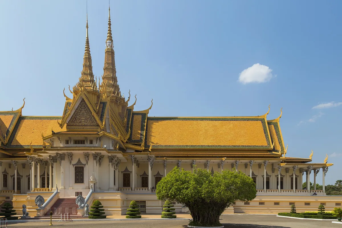 Forsiden af Tronhallen i Det Kongelige Palads i Phnom Penh, en korsformet bygning med gyldne tage og tvillingespir, et klippet træ i forgrunden.